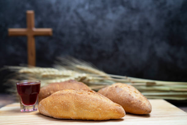 Communion consisting of a small glass of wine and loaves of bread on a table. A bushel of wheat and wooden cross in background