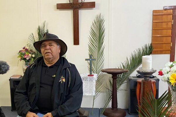 Rev Robert Jetta sitting at the front of Waroona Uniting Church with palm fronds and cross in the background.
